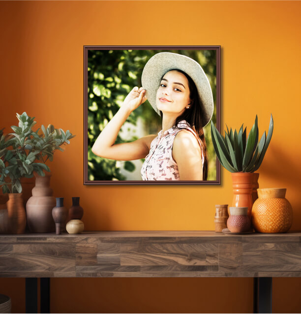Portrait of a young woman in a sunhat smiling at the camera, displayed in a square frame on an orange wall above a wooden console with decorative vases and plants.